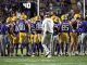 LSU football players in purple and gold snacks gathering in a huddle on the field as a coach walks through center.