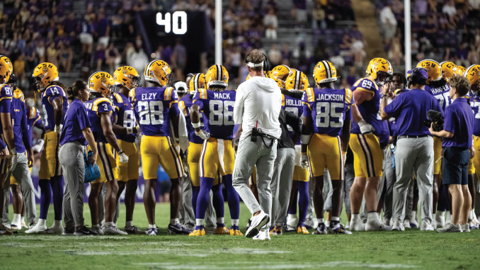 LSU football players in purple and gold snacks gathering in a huddle on the field as a coach walks through center.