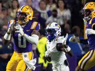LSU players in purple and gold chase a football; a teammate (#0) catches a pass as a defender in white closes in and another LSU player (#2) runs alongside.