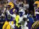 LSU players in purple and gold chase a football; a teammate (#0) catches a pass as a defender in white closes in and another LSU player (#2) runs alongside.