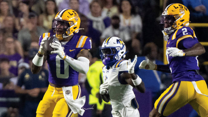 LSU players in purple and gold chase a football; a teammate (#0) catches a pass as a defender in white closes in and another LSU player (#2) runs alongside.