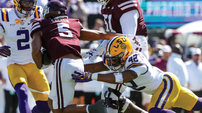 LSUFBallVMissState101726_DahvonKeys_JMailhes | TigerRag.com LSU defender in a white uniform (#42) reaches to tackle a maroon-clad opponent as teammates dive into a crowded pile during a college football game.