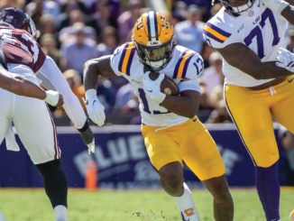 LSU running back in white and gold uniform carries the ball while defenders in maroon pursue during a college football game on a sunny day.