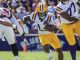LSU running back in white and gold uniform carries the ball while defenders in maroon pursue during a college football game on a sunny day.