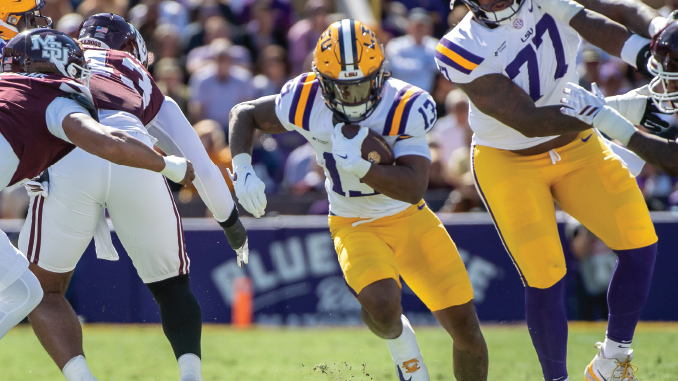LSU running back in white and gold uniform carries the ball while defenders in maroon pursue during a college football game on a sunny day.