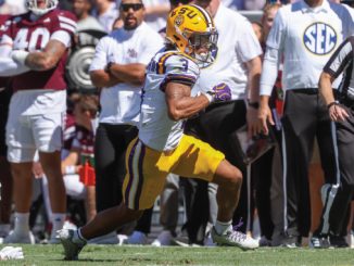 LSU running back in purple-yellow uniform carries ball past a fallen defender on a sunny sideline during a college football game; SEC official and onlookers in background.