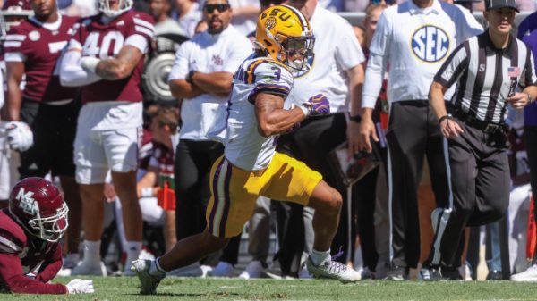 LSU running back in purple-yellow uniform carries ball past a fallen defender on a sunny sideline during a college football game; SEC official and onlookers in background.
