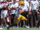 LSU running back in purple-yellow uniform carries ball past a fallen defender on a sunny sideline during a college football game; SEC official and onlookers in background.