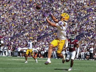 LSU football player in white jersey and yellow pants jumps to catch a pass as defenders pursue, packed purple-gold stadium in the background