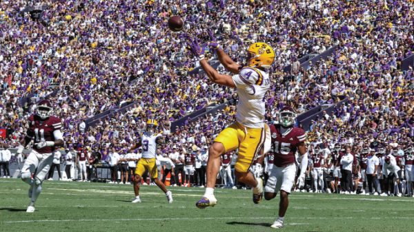 LSU football player in white jersey and yellow pants jumps to catch a pass as defenders pursue, packed purple-gold stadium in the background