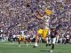 LSU football player in white jersey and yellow pants jumps to catch a pass as defenders pursue, packed purple-gold stadium in the background