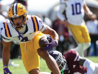 LSU running back in white and purple uniform carries a football while dodging a tackler on a grassy field.