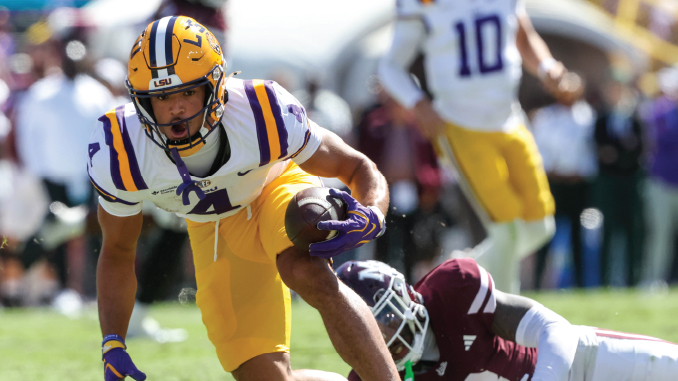 LSU running back in white and purple uniform carries a football while dodging a tackler on a grassy field.