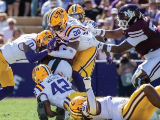Players in yellow-helmeted LSU uniforms tackle a maroon-clad opponent as a pileup forms on the field during a sunny game.