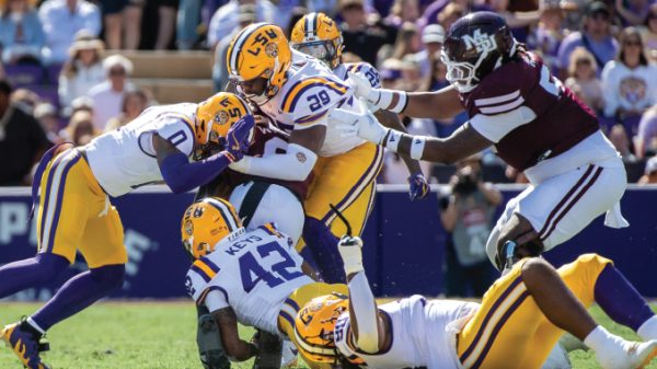 Players in yellow-helmeted LSU uniforms tackle a maroon-clad opponent as a pileup forms on the field during a sunny game.