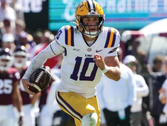 LSU football quarterback wearing #10 in white and gold uniform runs with ball during a game, crowd in background