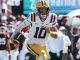 LSU football quarterback wearing #10 in white and gold uniform runs with ball during a game, crowd in background