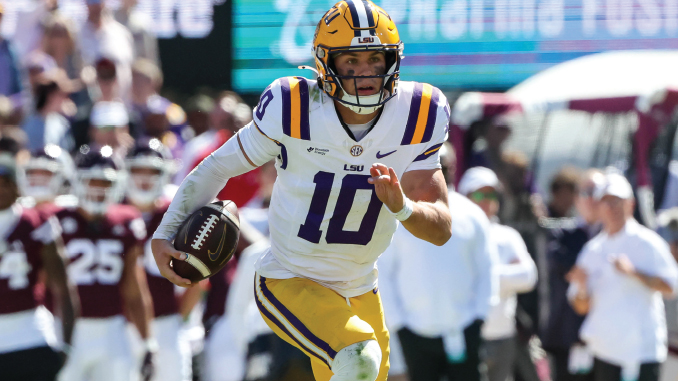 LSU football quarterback wearing #10 in white and gold uniform runs with ball during a game, crowd in background
