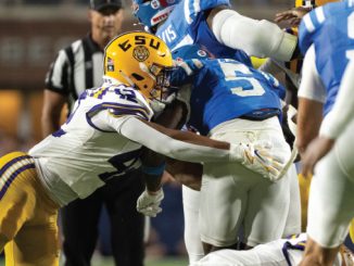 Football tackle during a college game: a LSU player in white-gold helmet grips an opposing blue-jersey runner in the open field near the end zone line behind, referee in background.