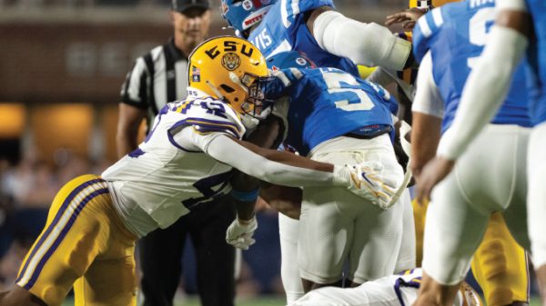 Football tackle during a college game: a LSU player in white-gold helmet grips an opposing blue-jersey runner in the open field near the end zone line behind, referee in background.