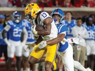 LSU running back wearing number 13 carries the football as a defender in blue tackles from behind during a college football game in progress on the field.