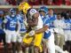 LSU running back wearing number 13 carries the football as a defender in blue tackles from behind during a college football game in progress on the field.