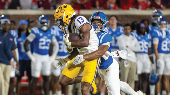LSU running back wearing number 13 carries the football as a defender in blue tackles from behind during a college football game in progress on the field.