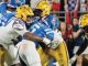 LSU players in yellow helmets tackle a blue-clad opponent at the line of scrimmage with fans in the background.