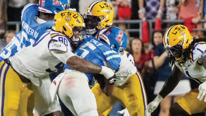 LSU players in yellow helmets tackle a blue-clad opponent at the line of scrimmage with fans in the background.