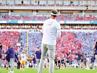 Coach standing on a football field facing a packed stadium during a game-day moment.