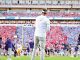 Coach standing on a football field facing a packed stadium during a game-day moment.