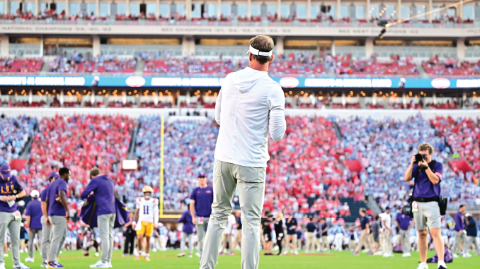 Coach standing on a football field facing a packed stadium during a game-day moment.