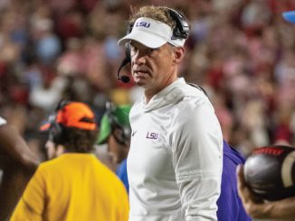 LSU football coach in a white polo and headset on the sideline during a game, crowd blurred in the background.