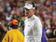 LSU football coach in a white polo and headset on the sideline during a game, crowd blurred in the background.