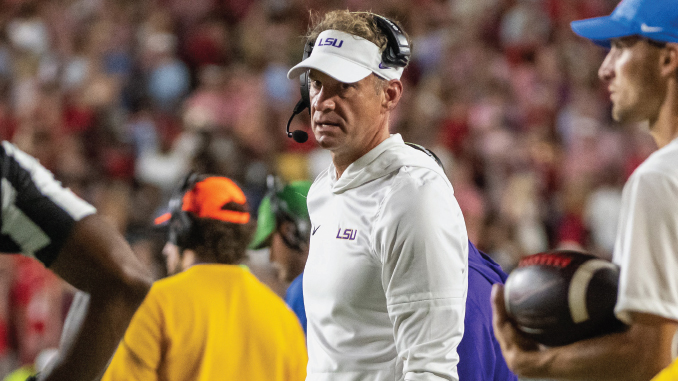 LSU football coach in a white polo and headset on the sideline during a game, crowd blurred in the background.