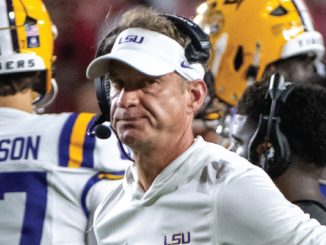 LSU football coach with a white cap and headset on the sideline, players in purple and gold uniforms nearby.