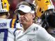 LSU football coach with a white cap and headset on the sideline, players in purple and gold uniforms nearby.