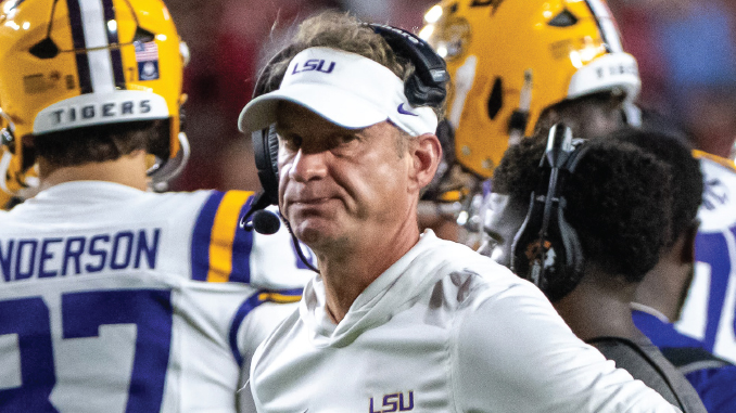 LSU football coach with a white cap and headset on the sideline, players in purple and gold uniforms nearby.