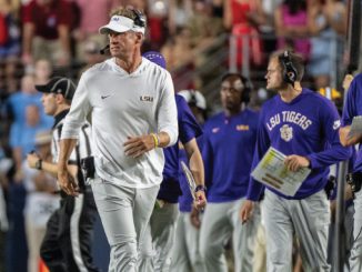 LSU coach in white shirt and visor walking on the sideline during a college football game, with staff around him and fans in the stands behind.