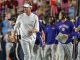 LSU coach in white shirt and visor walking on the sideline during a college football game, with staff around him and fans in the stands behind.