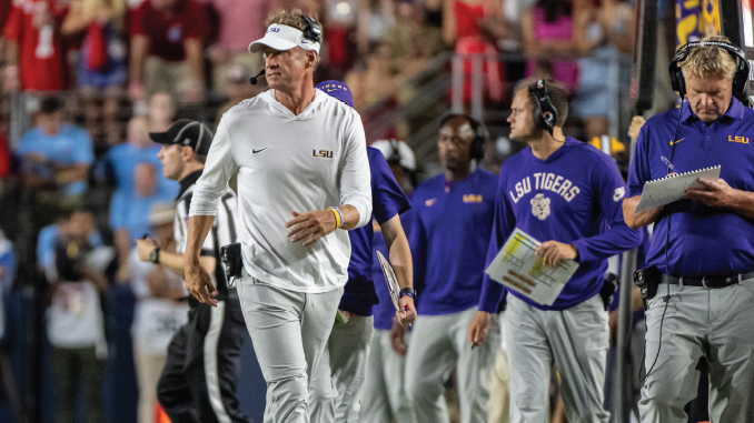 LSU coach in white shirt and visor walking on the sideline during a college football game, with staff around him and fans in the stands behind.