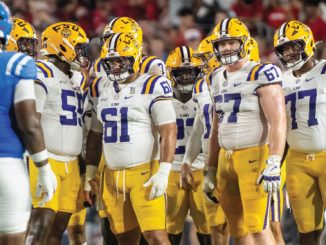 LSU football players in white jerseys and gold pants huddle on the field, facing blue-clad opponents.