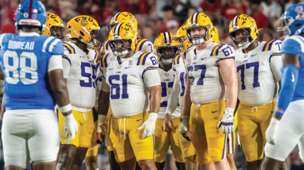 LSU football players in white jerseys and gold pants huddle on the field, facing blue-clad opponents.