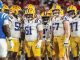 LSU football players in white jerseys and gold pants huddle on the field, facing blue-clad opponents.