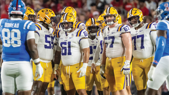 LSU football players in white jerseys and gold pants huddle on the field, facing blue-clad opponents.