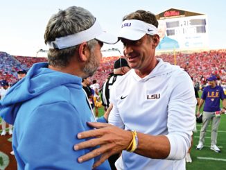 Two men on a college football field share a friendly hug as the packed stadium looks on in the background.