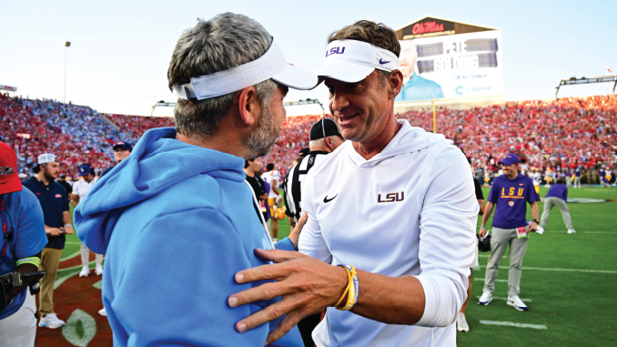 Two men on a college football field share a friendly hug as the packed stadium looks on in the background.