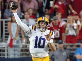 LSU quarterback wearing number 10 throws a pass on the football field with fans in the stands behind him.