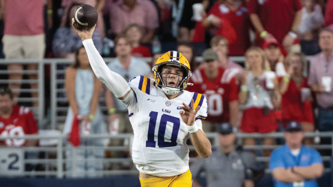 LSU quarterback wearing number 10 throws a pass on the football field with fans in the stands behind him.
