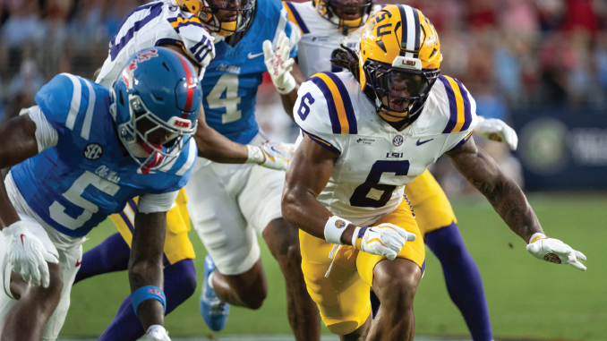 LSU Tigers running back wearing number 6 sprints with the ball, chased by blue-clad defenders during a game on the field.